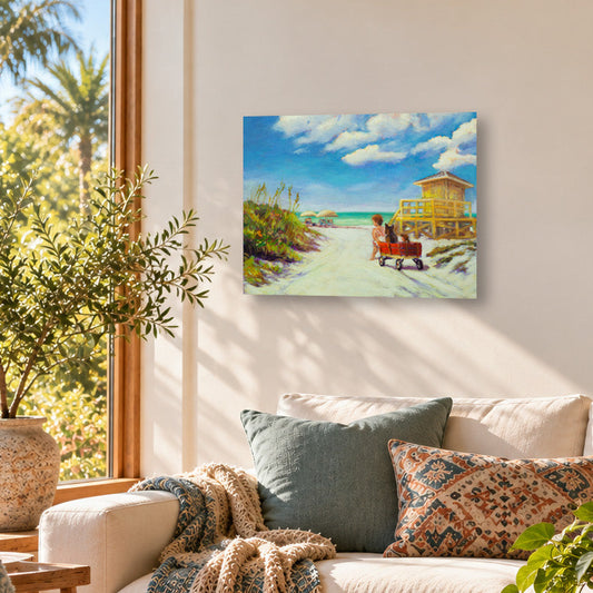 Colorful beach art print featuring a child pulling a red wagon along a sandy path toward the ocean, with a yellow lifeguard tower and bright blue sky, displayed above a cream sofa in a sunlit coastal living room with natural wood furniture.