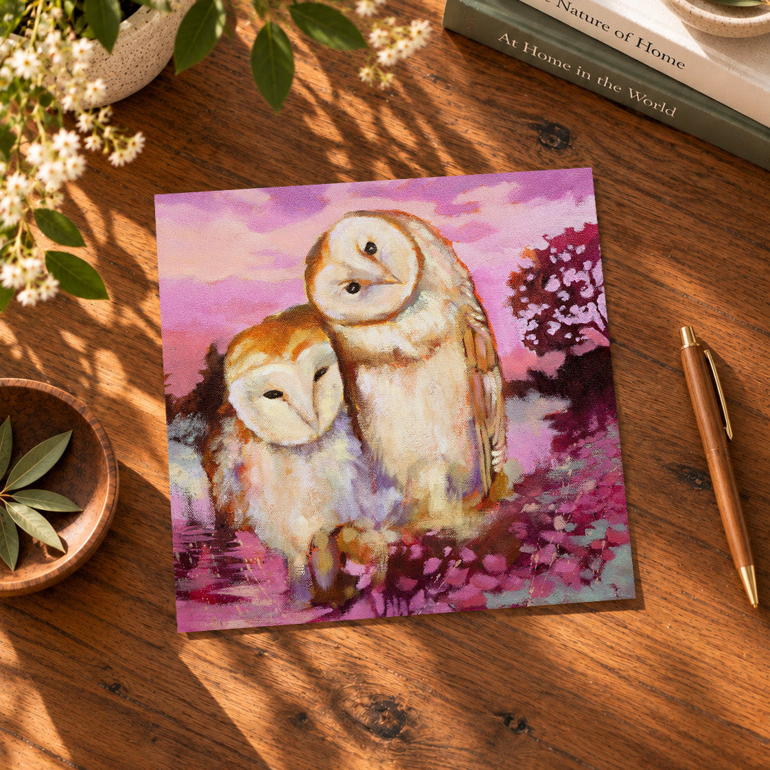 Quiet Support square barn owl art print displayed on a sunlit wooden table, featuring two owls leaning together in a colorful pink and purple landscape.