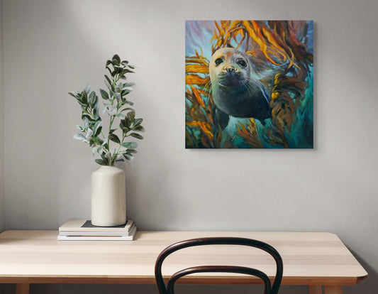 Painting of a young seal swimming through a glowing kelp forest, titled “Wild in the Weeds,” hanging on a light gray wall above a wooden desk with a vase and books.