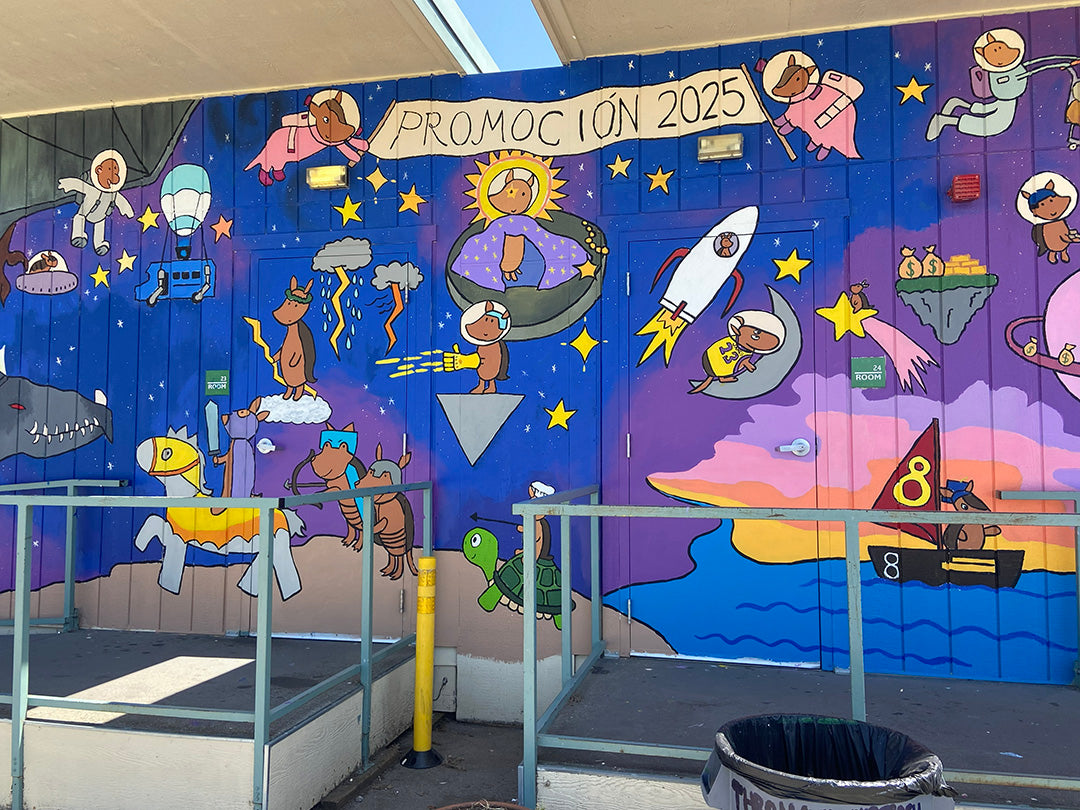 A colorful outdoor mural painted on blue shipping containers under a patio roof. The mural features playful cartoon astronauts, a rocket ship, stars, planets, and a banner reading 'PROMOCION 2025'.