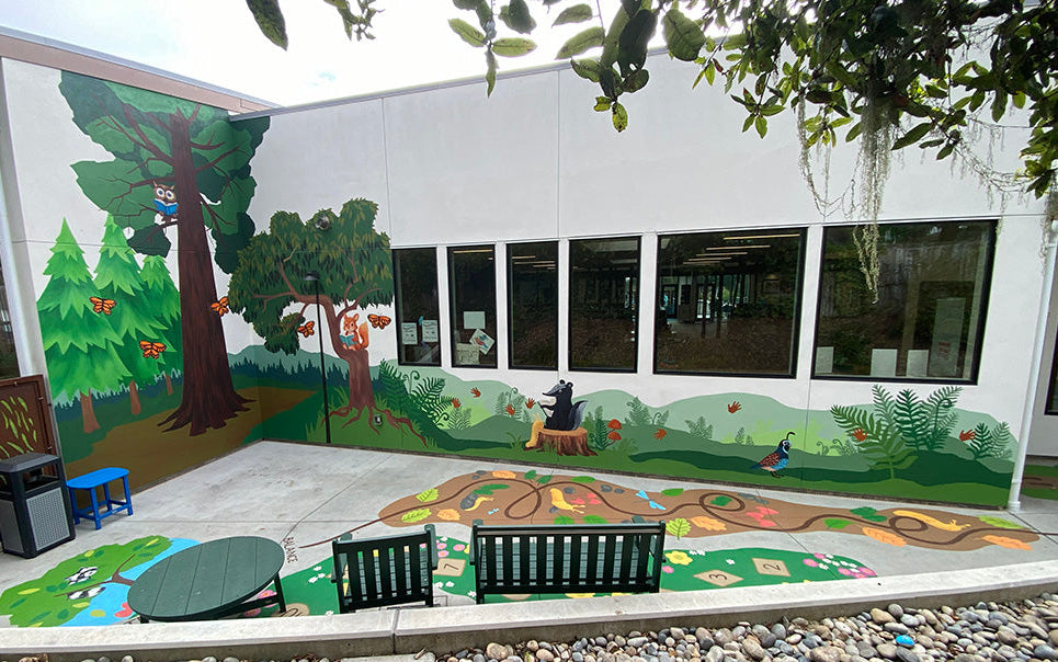 Outdoor courtyard wall mural painted with forest trees, animals, and a winding trail, shown beside a white building with large windows. Benches, a small table, rocks, and hanging leaves are visible in the foreground.