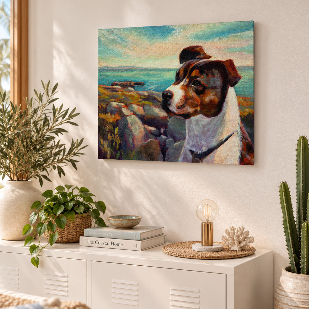 Colorful oil painting of a brown-and-white dog overlooking a rocky ocean coastline, displayed above a white cabinet in a sunlit coastal-style living room with natural textures, greenery, and warm neutral decor.