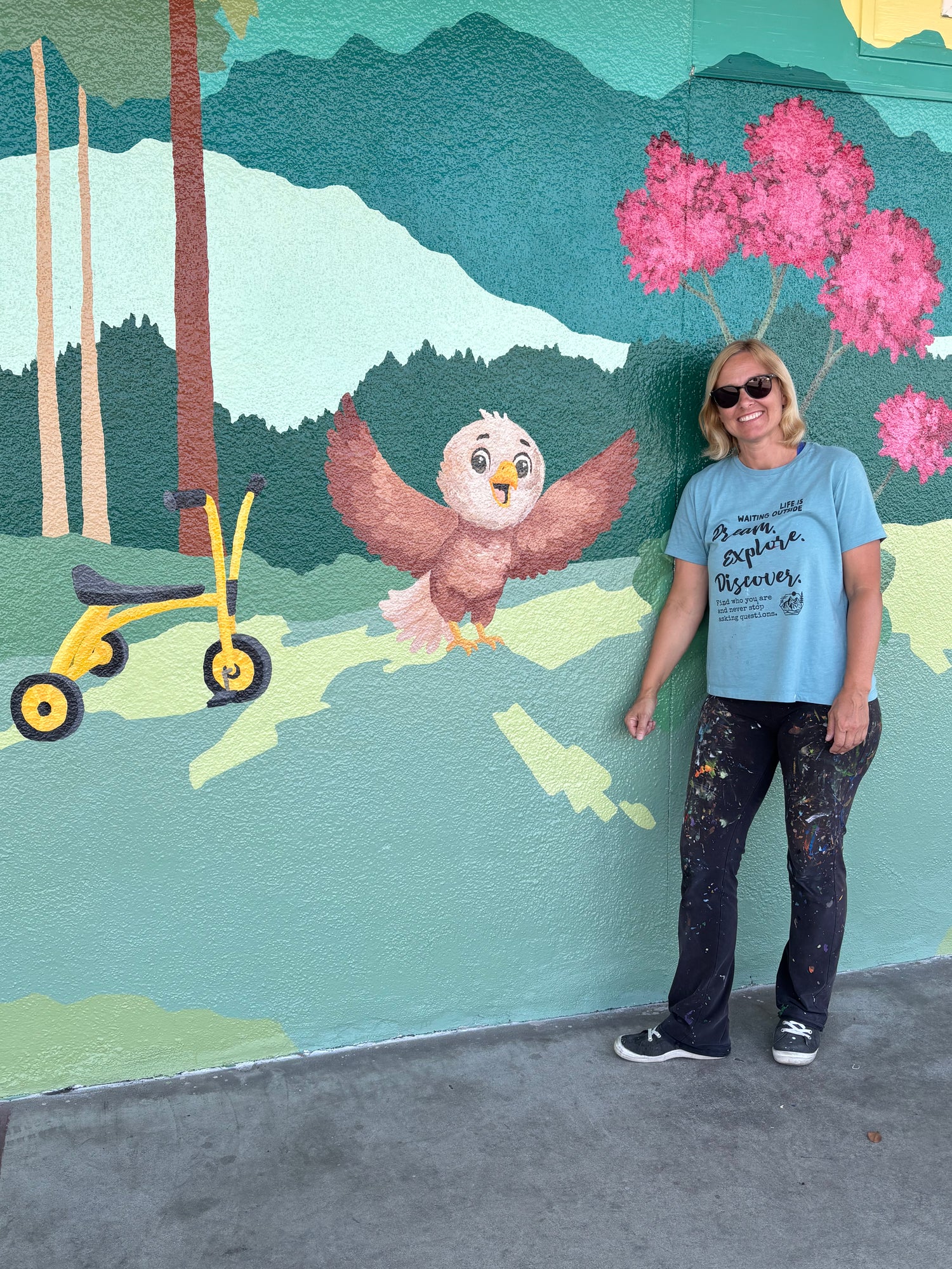 A woman wearing a light blue graphic t-shirt and paint-splattered jeans stands in front of a colorful mural depicting a bird, pink trees, a yellow tricycle, and mountains.