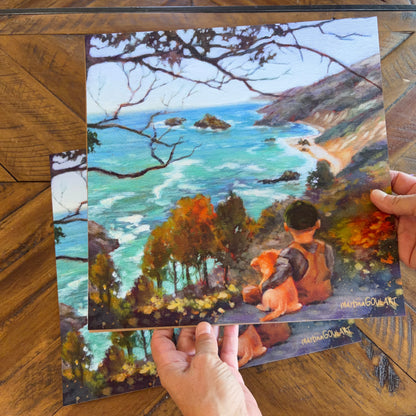 Hands holding a print of a boy with a dog looking out to the Pacific Ocean near Big Sur. 