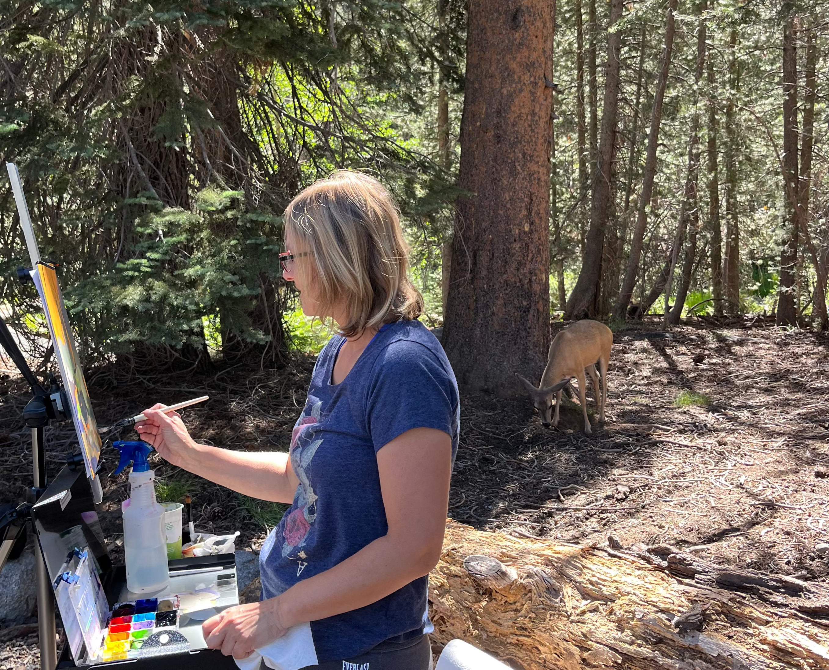 Person in a blue shirt painting outdoors in a forest setting, deer behind her, sunny day, brown color forest in the background.