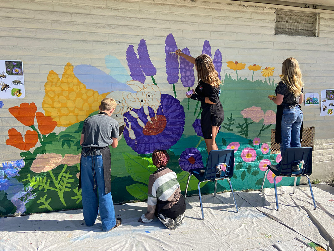 Several people painting a colorful mural on an outdoor brick wall, featuring flowers, leaves, and a rabbit. Two people are standing on chairs, and one person is kneeling in the foreground on a white drop cloth.