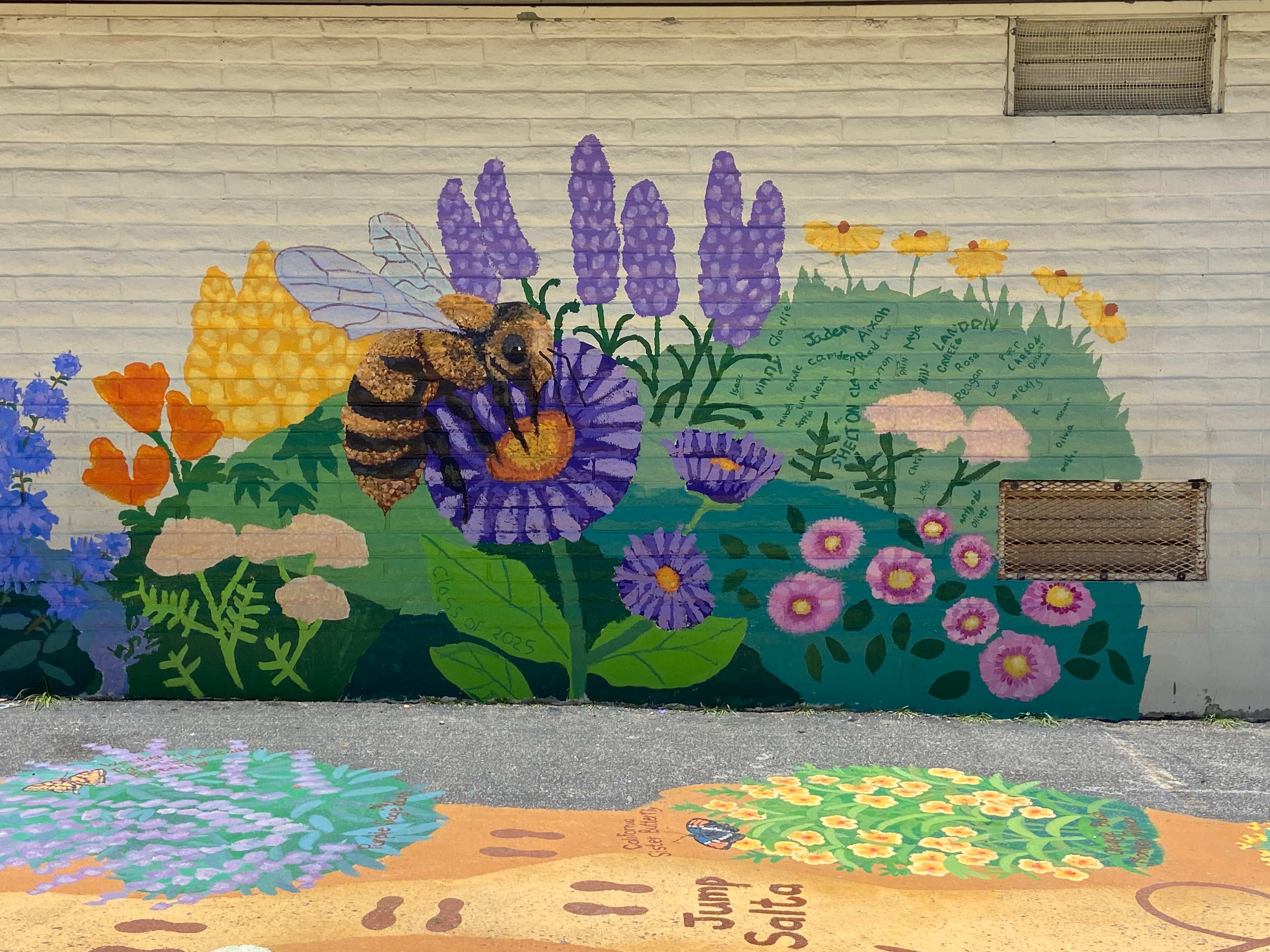 A colorful mural painted on a white brick wall featuring a bee among purple flowers, green leaves, yellow blossoms, and pink and orange flowers. A paved walkway with painted sections is visible in the foreground.