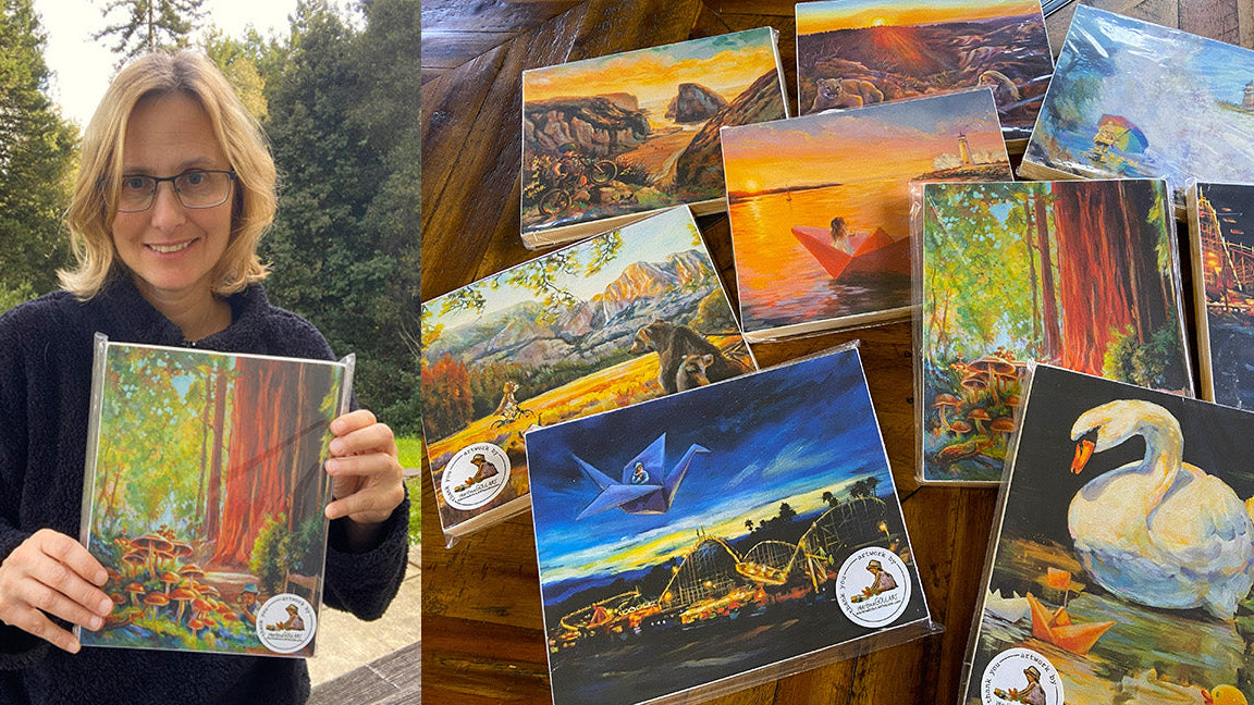 Artist Martina Goulart holding a painting next to a collection of fine art prints featuring landscapes and animals.