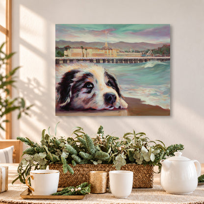 Colorful oil painting of a black-and-white puppy resting on the beach with the Santa Cruz Beach Boardwalk and ocean waves in the background, displayed above a wooden dining table in a warm, sunlit coastal home with natural greenery and cozy neutral decor.