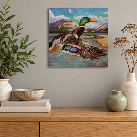 Original wildlife oil painting of a mallard duck flying over a mountain lake, with colorful blue and green feathers, by Martina Goulart, displayed above a wooden cabinet in a modern living room.