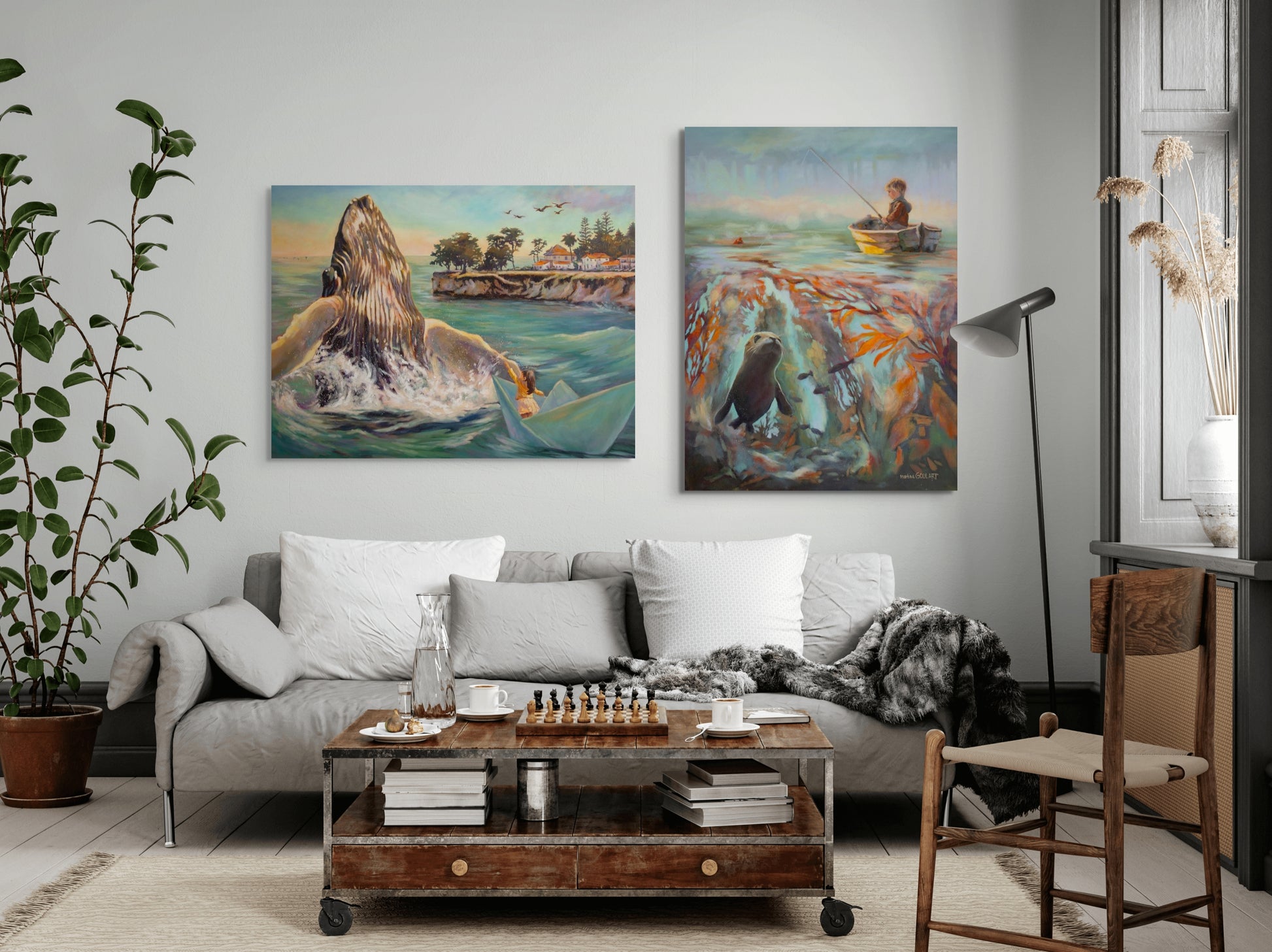 Two large coastal paintings hanging above a sofa: one of a girl in a paper boat beside a breaching whale, and the other of a boy fishing above a kelp forest with a seal.