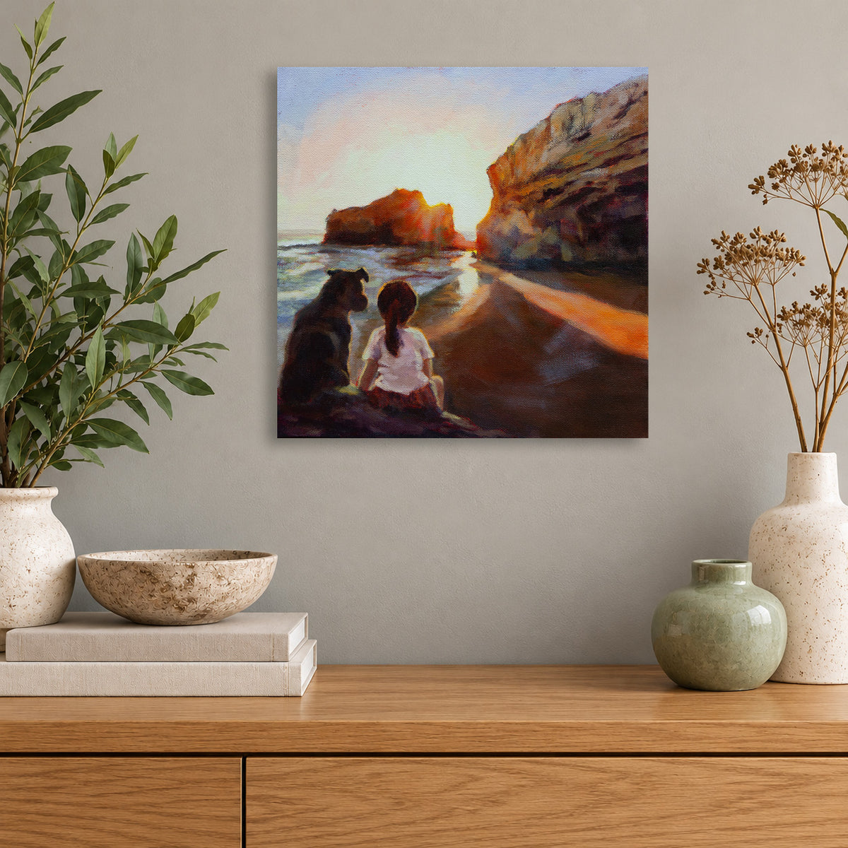 Art print by Martina Goulart featuring a young girl and her dog watching the sunset along a rocky California beach, displayed above a natural wood console in a coastal-inspired room.