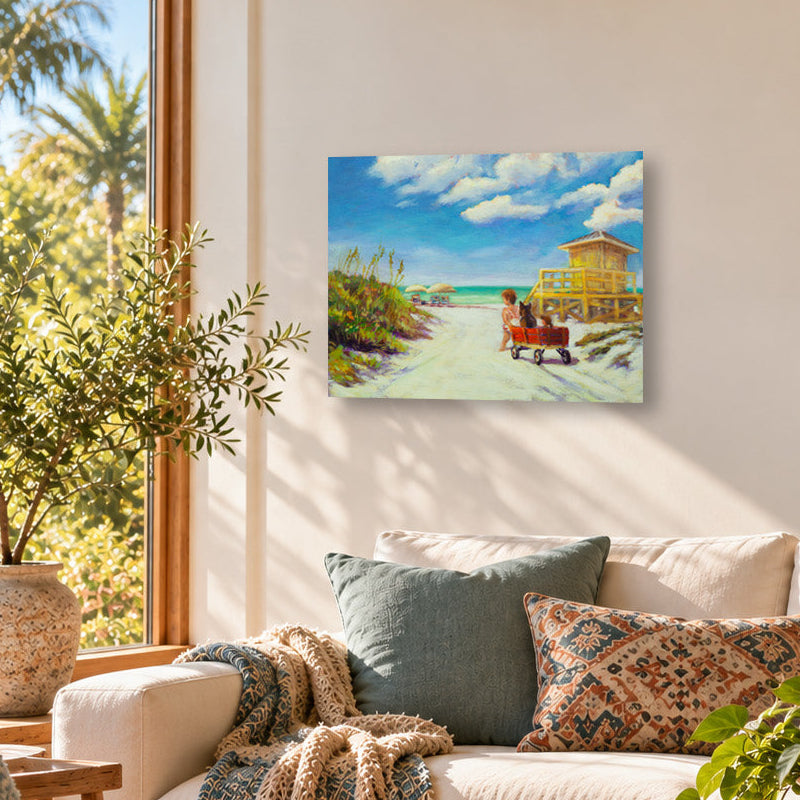 Colorful beach art print featuring a child pulling a red wagon along a sandy path toward the ocean, with a yellow lifeguard tower and bright blue sky, displayed above a cream sofa in a sunlit coastal living room with natural wood furniture.