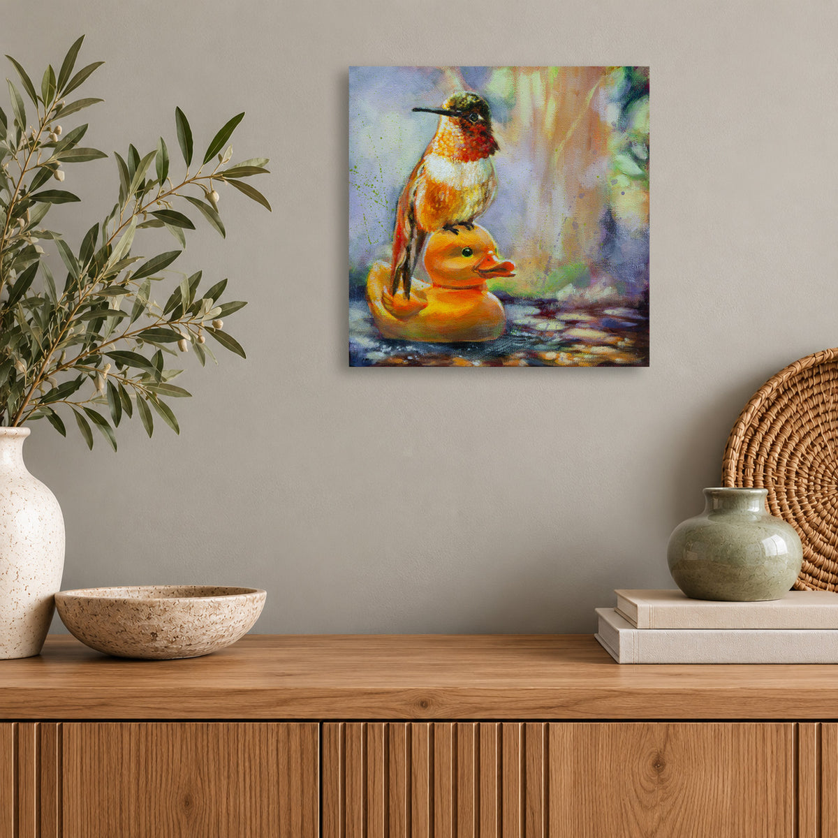 Whimsical oil painting of a colorful hummingbird perched on a yellow rubber duck floating in water, displayed above a natural wood console in a modern home with olive branches, woven accents, and neutral decor.