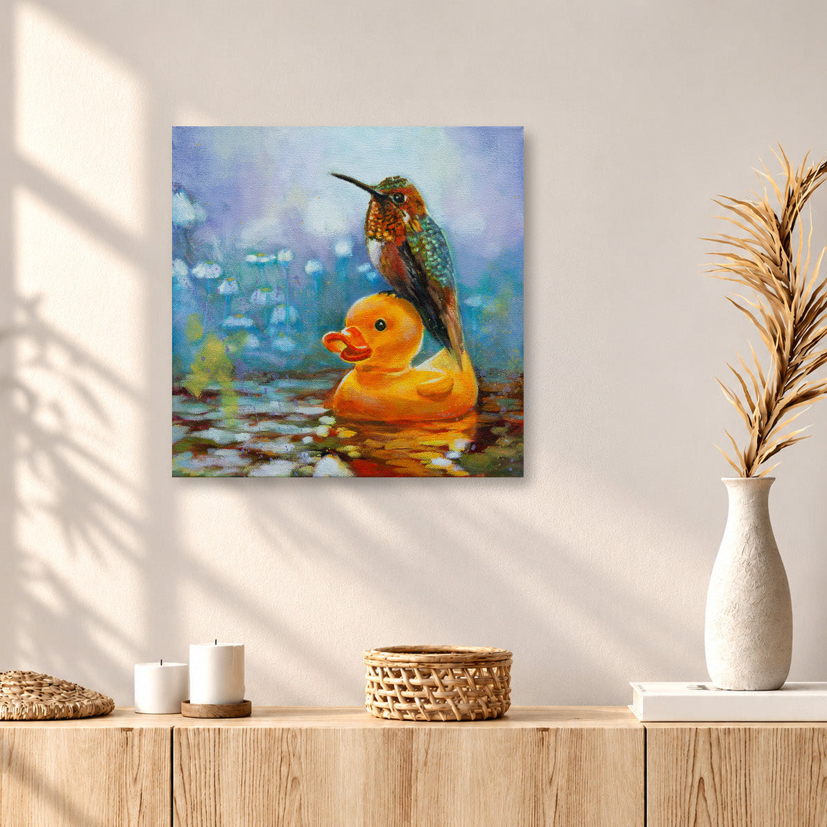 Colorful oil painting of a hummingbird perched on a bright yellow rubber duck floating in water, displayed above a light oak cabinet in a warm, sunlit modern home with soft window shadows, woven textures, and a ceramic vase.