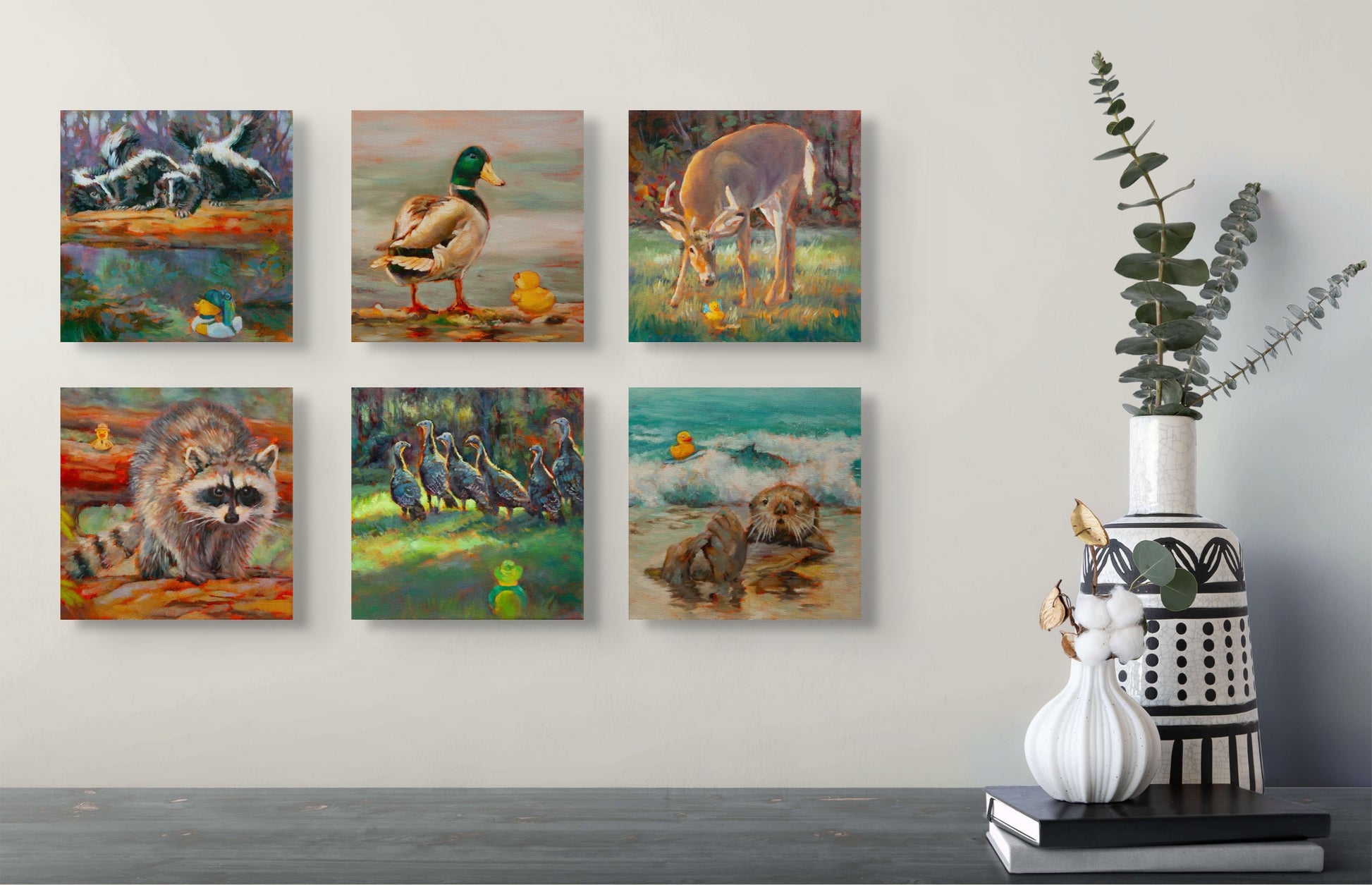 Six Wild Neighbors paintings — raccoons, mallard, deer, turkeys, and otter — hanging above a gray table with white and black vases and a green plant.