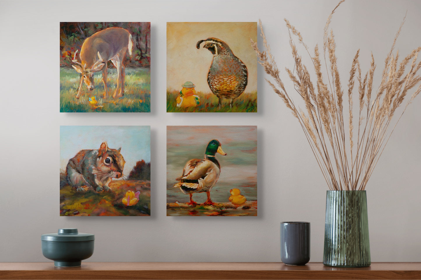 Four Wild Neighbors paintings — deer, quail, squirrel, and mallard — hanging above a brown table with a green vase, dry grass, and two gray cups.