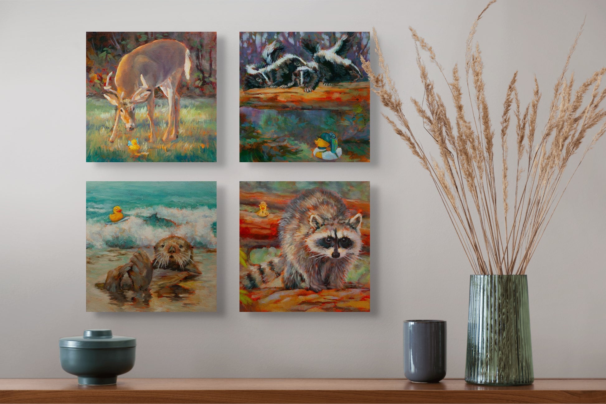 Four Wild Neighbors paintings — deer, skunks, otter, and raccoon — hanging above a brown table with a green vase, dry grass, and two gray cups.