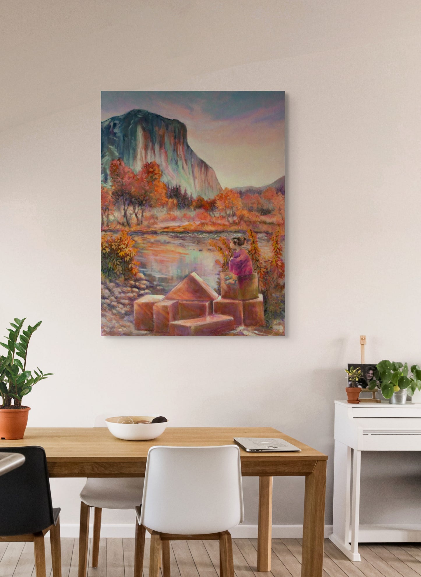 Painting One Day, El Cap. hanging in a dining room with tan table, white chairs, bowl on table, and green houseplants, warm inviting setting.