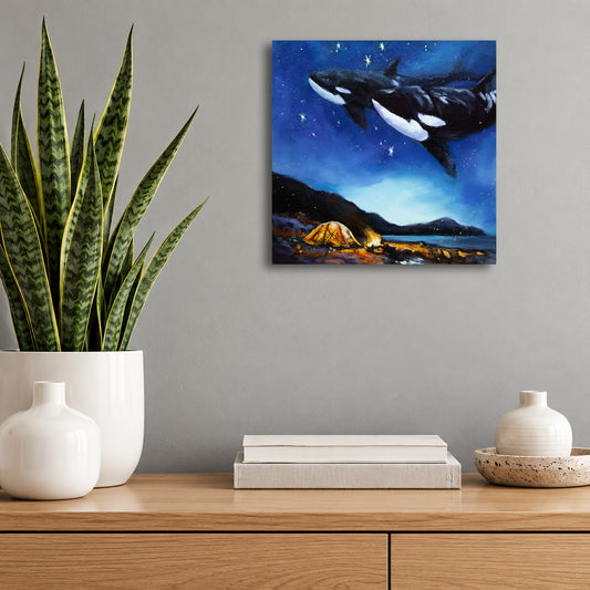 Original oil painting by Martina Goulart featuring two orcas swimming through a magical starry night sky above a glowing campsite by the ocean, displayed on a gray wall above a wooden cabinet.