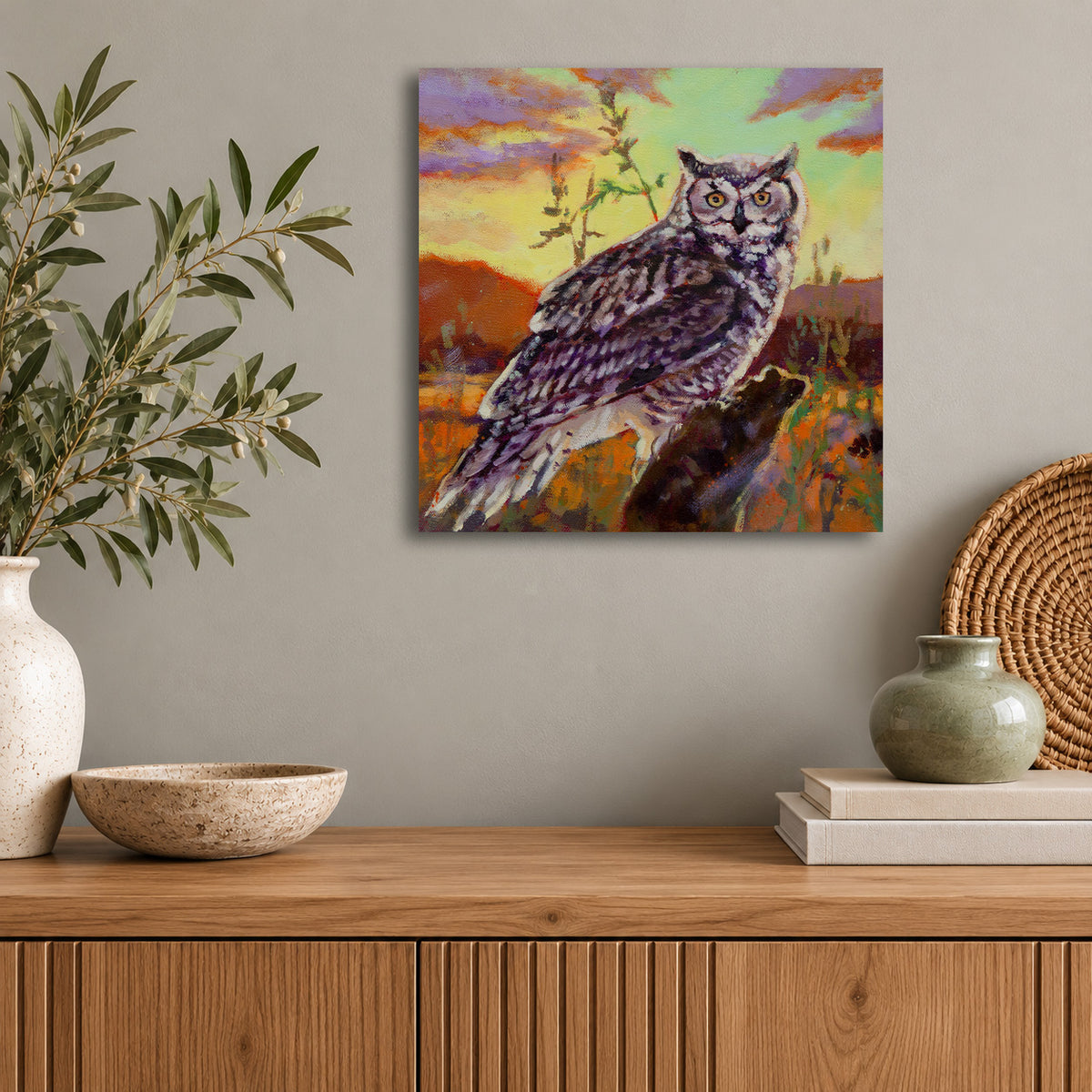 Original wildlife oil painting of a great horned owl perched on a branch against a colorful sunset sky by Martina Goulart, displayed in a modern living room with natural wood furnishings.
