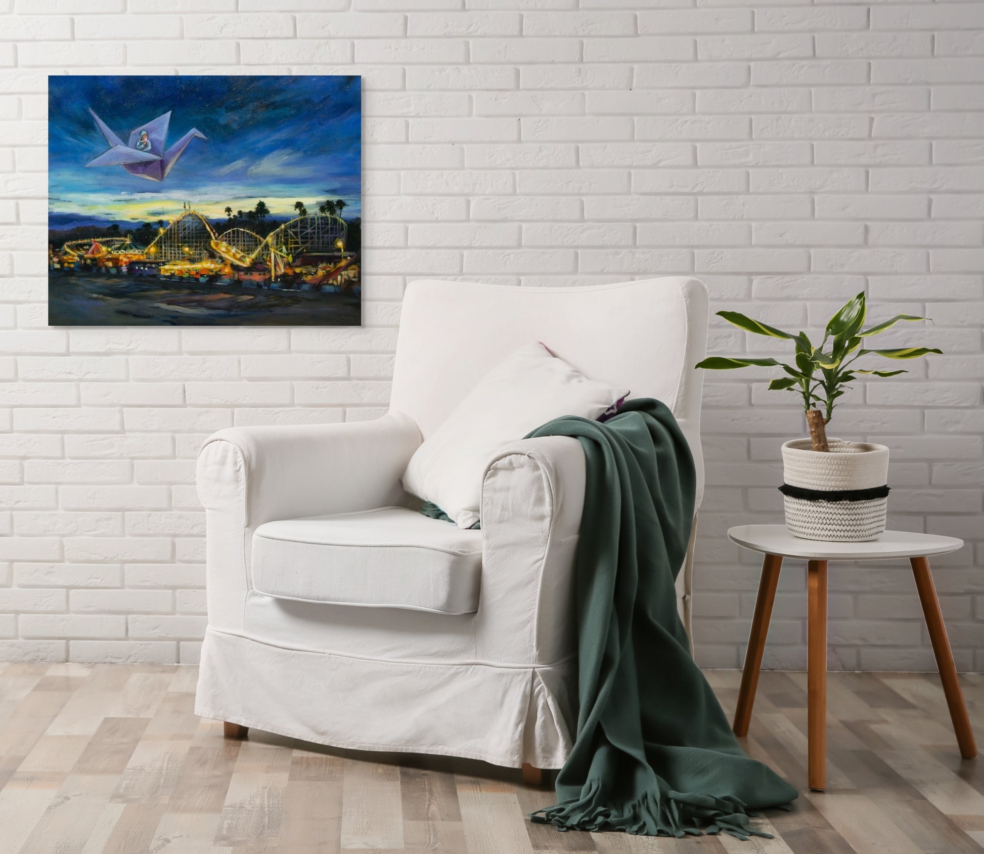 White armchair with green blanket and an original painting of the Santa Cruz Beach Boardwalk at night, with blue-purple skies and glowing orange lights.