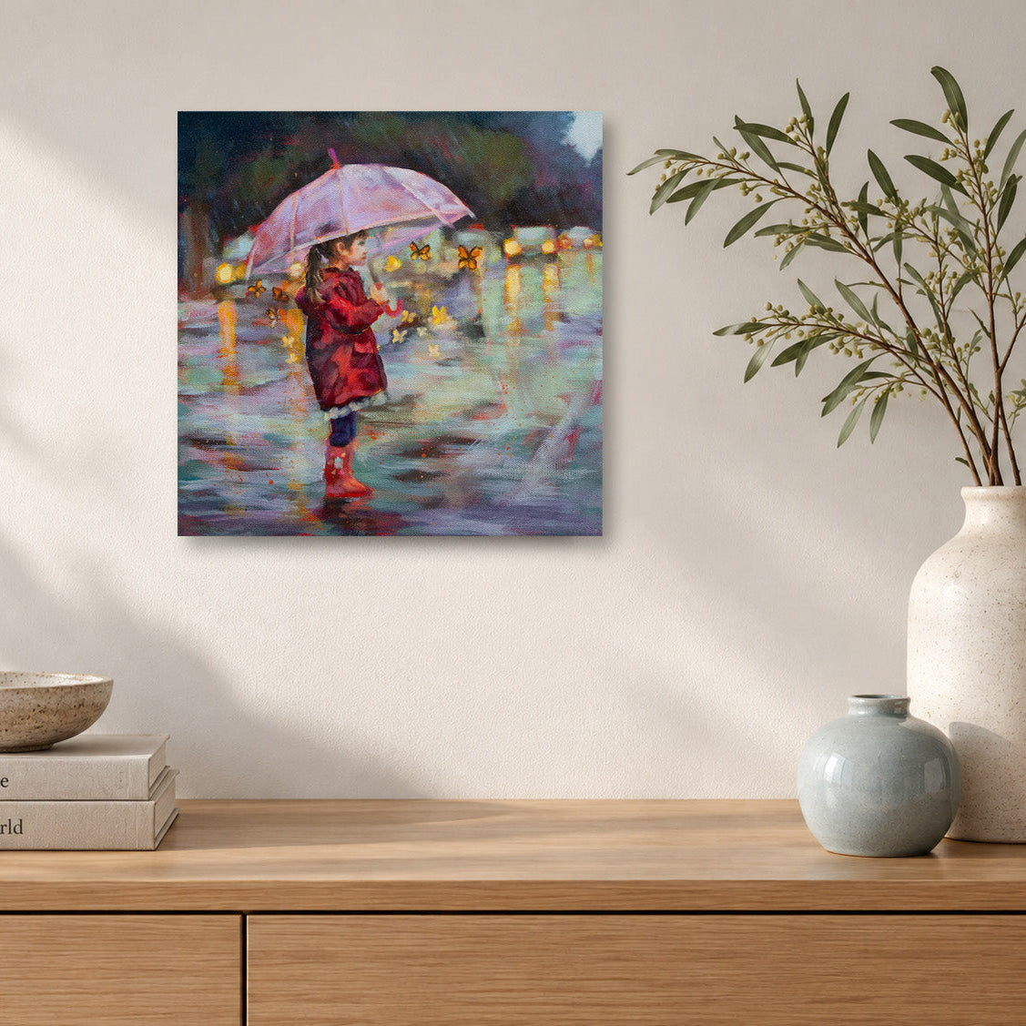 alt text pleas;e
Colorful painting by Martina Goulart of a young girl in a red raincoat holding a pink umbrella, surrounded by butterflies on a rainy street, displayed above a natural wood cabinet in a softly lit modern living room with neutral decor and ceramic vases.