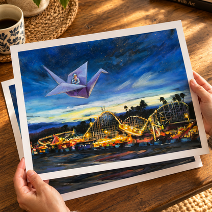  Hands holding an art print of Voyage of the Paper Crane over a warm wooden table. The artwork shows a boy flying on a purple paper crane above the glowing Santa Cruz Beach Boardwalk at night. 