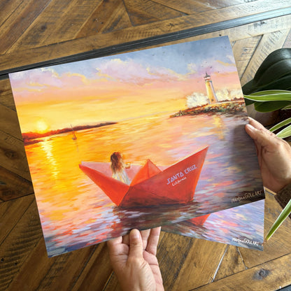 Hands holding an art print of Safe Harbor over a warm wooden table. The artwork shows a girl floating in a red paper boat near Santa Cruz harbor and lighthouse. 