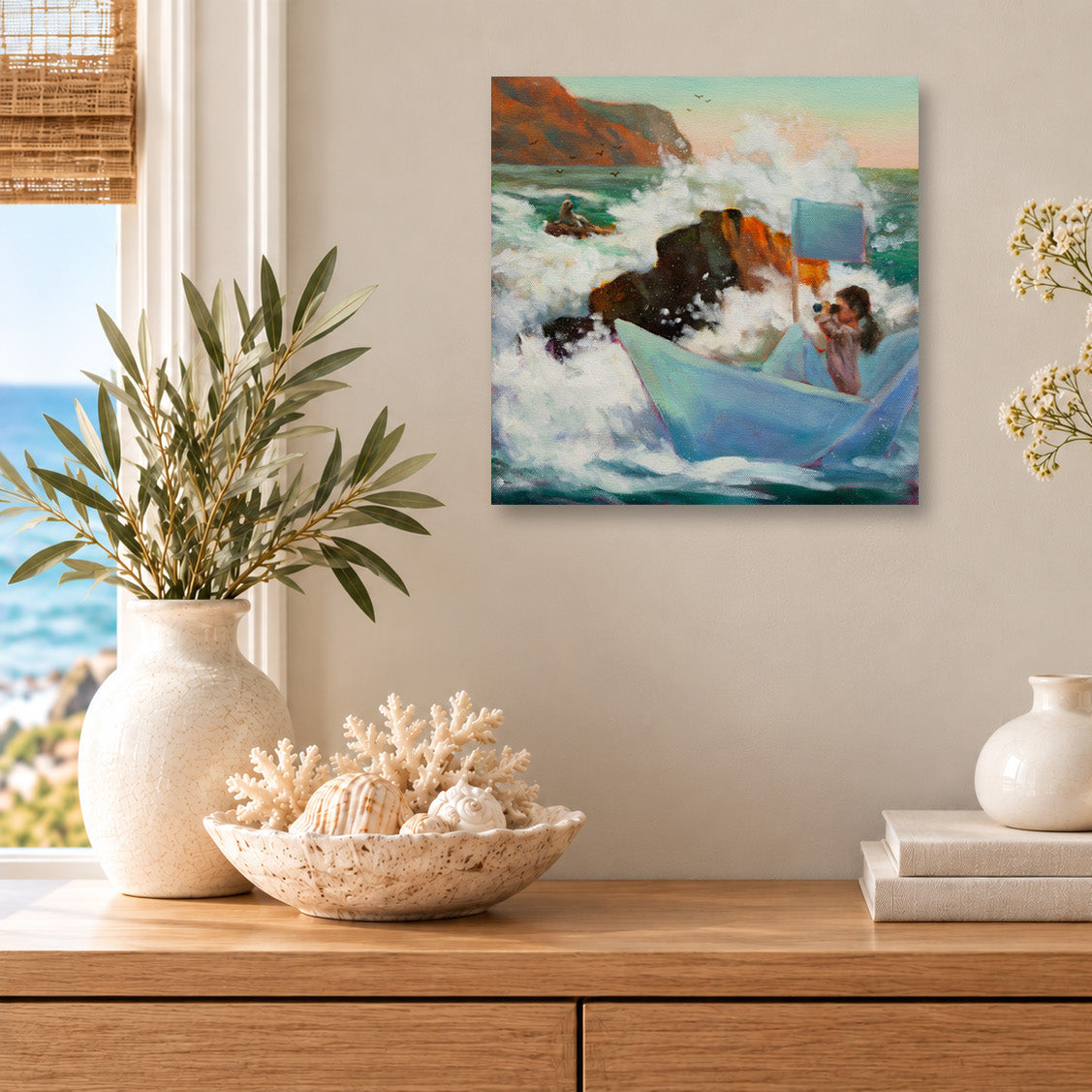 Whimsical coastal art print featuring a girl sailing a blue paper boat through crashing ocean waves, displayed above a wooden cabinet with seashells, books, and white ceramic vases in a sunlit beach-inspired room.