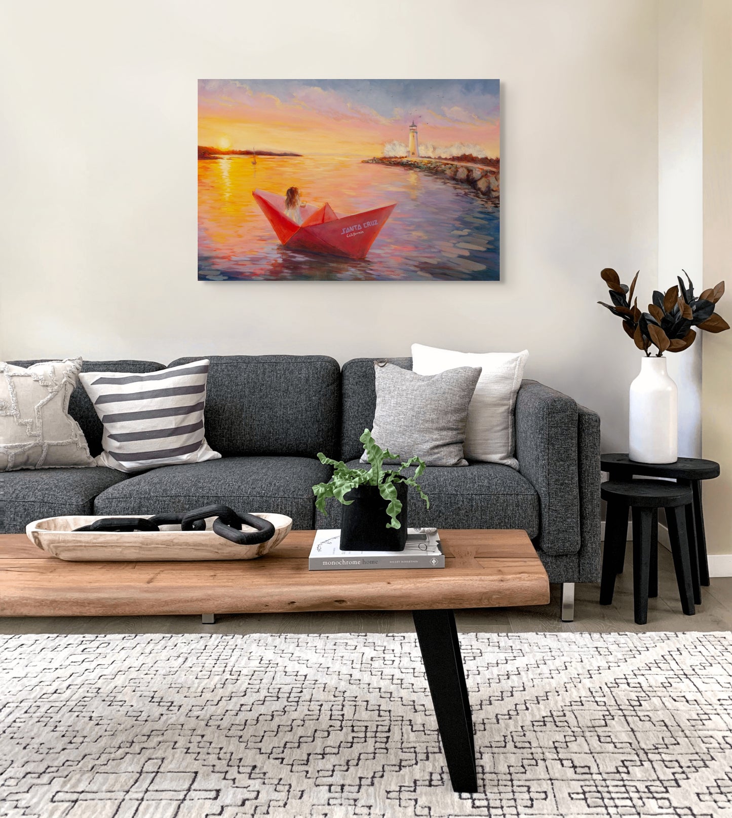 Living room with a gray sofa and wooden coffee table, featuring an original painting of a red boat at sunset with a lighthouse and paper boat entering Santa Cruz Harbor.