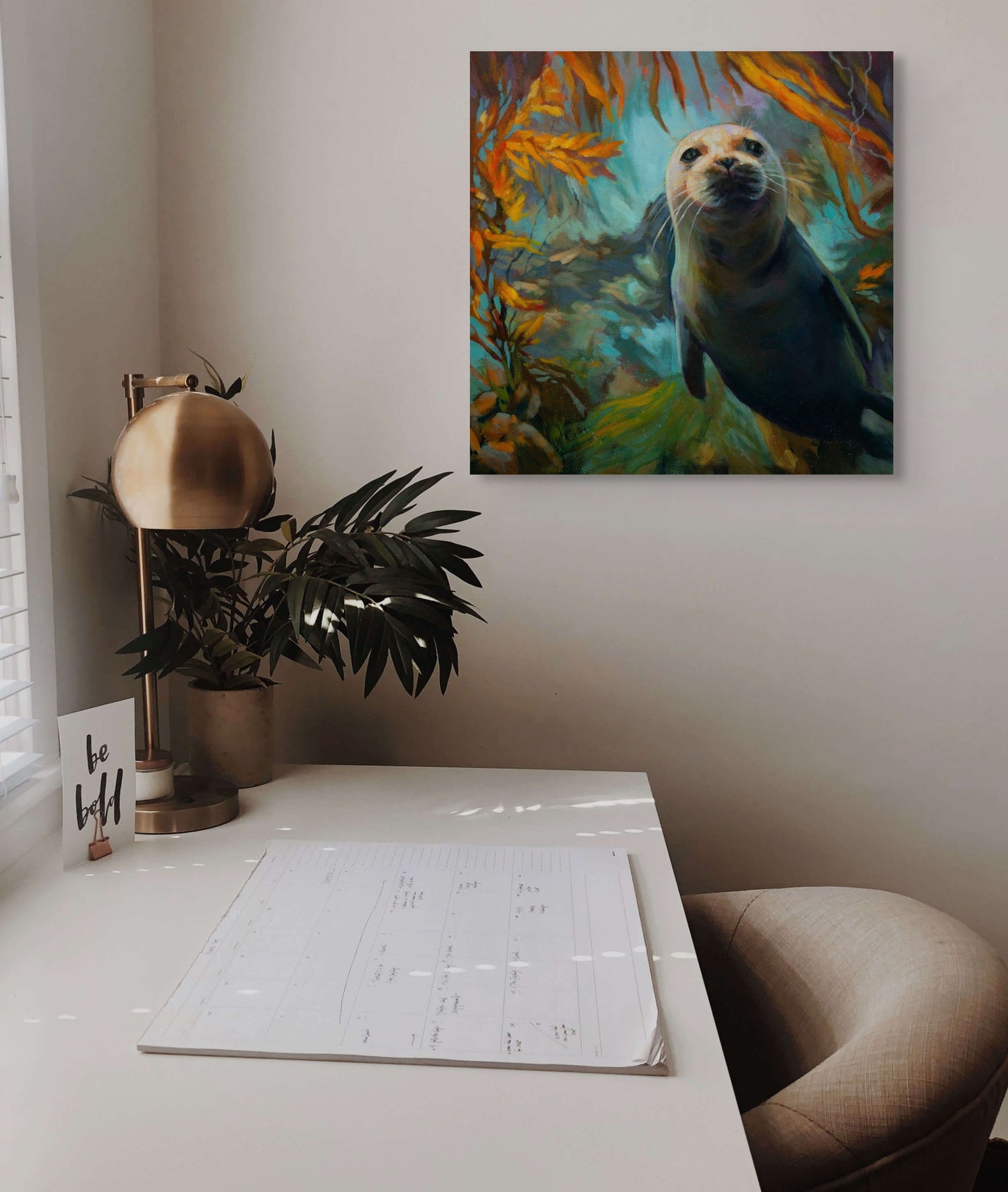 Painting of a young seal swimming upward through a glowing kelp forest, titled “Curious by Nature,” hanging on a light wall above a desk with plants and warm natural light.