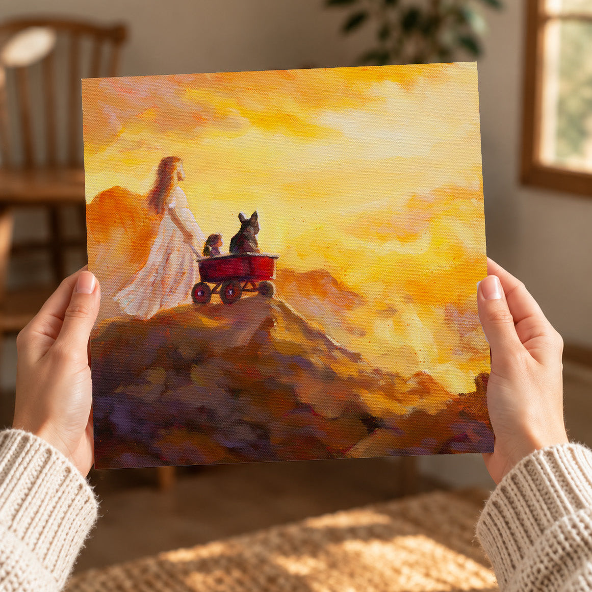 Hands holding a square art print of She Pulls the World Up, featuring a girl with a red wagon, dog, and doll overlooking a glowing mountain landscape.