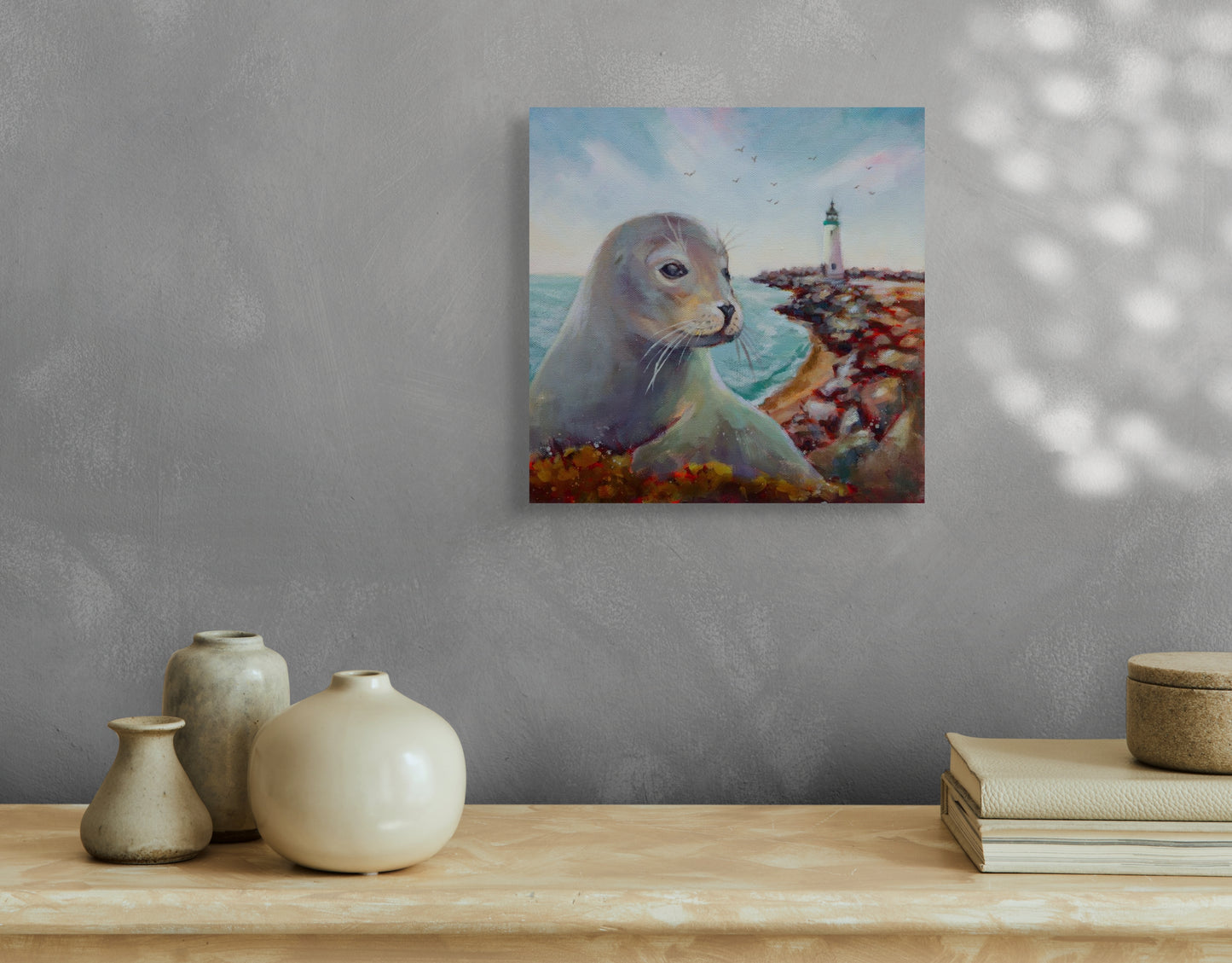 Painting of a seal by Walton Lighthouse hanging on a gray wall above a wooden shelf with ceramic vases and stacked books. Soft natural light falls across the wall.