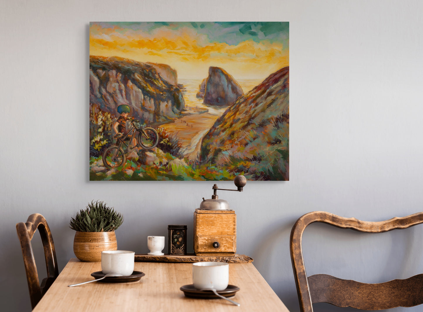 Painting Wild and Earned hanging above a natural wood table with two chairs and white teacups, warm orange-brown coastal artwork on the wall.