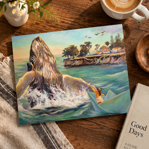 A square art print showing a dramatic painting of a whale rising from the ocean with a person in a small boat, styled on a wooden table with a coffee cup, flowers, a book, and a towel.